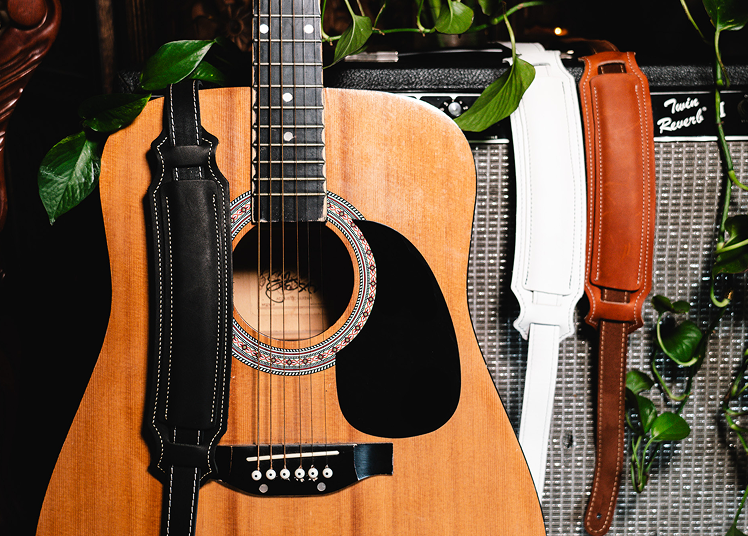 Acoustic Guitar with black leather strap draped over It leaning up against an AMP with two more leather straps displayed In white and brown.