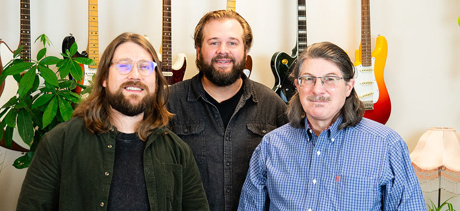 Greg Lancaster, Sr., Greg Lancaster II, and Charlton Moore from Franklin Strap standing In front of guitars hanging from a white wall.