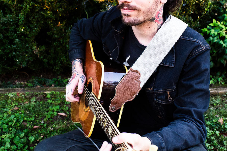 Man in denim shirt playing an accustic guitar sporting a white canvas guitar strap. 